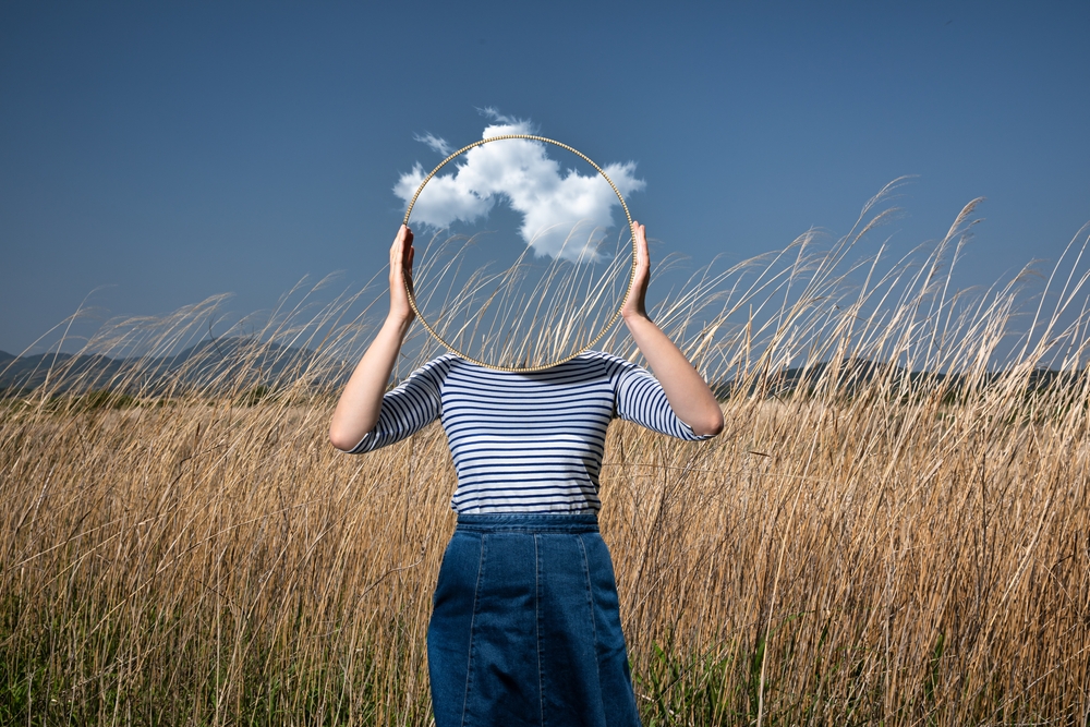 Person standing in field holding mirror in front of face with sky reflecting in it