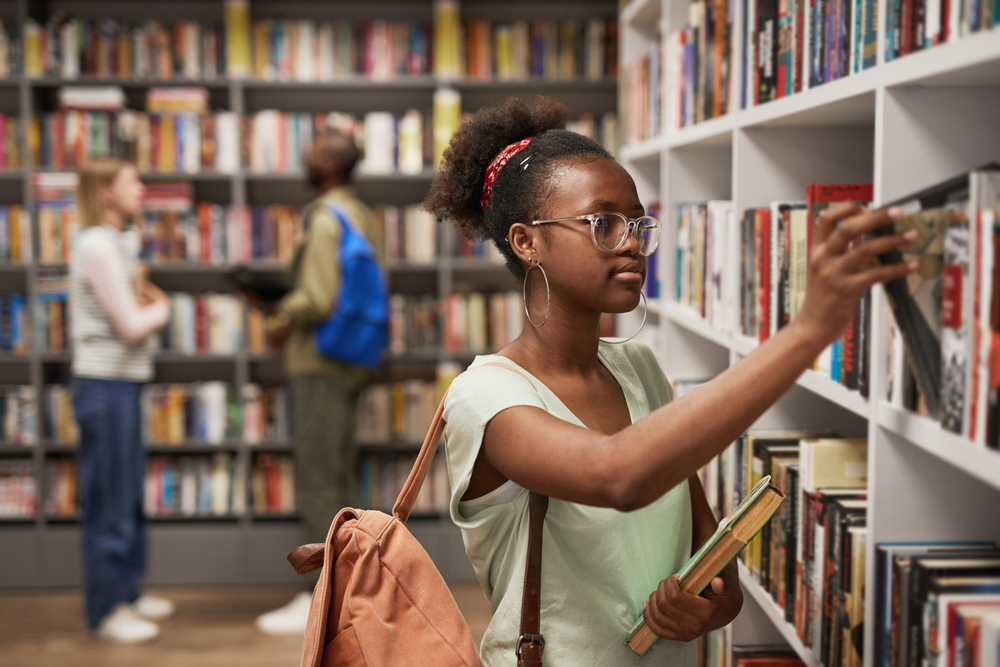 College student reaching for book on a library shelf.